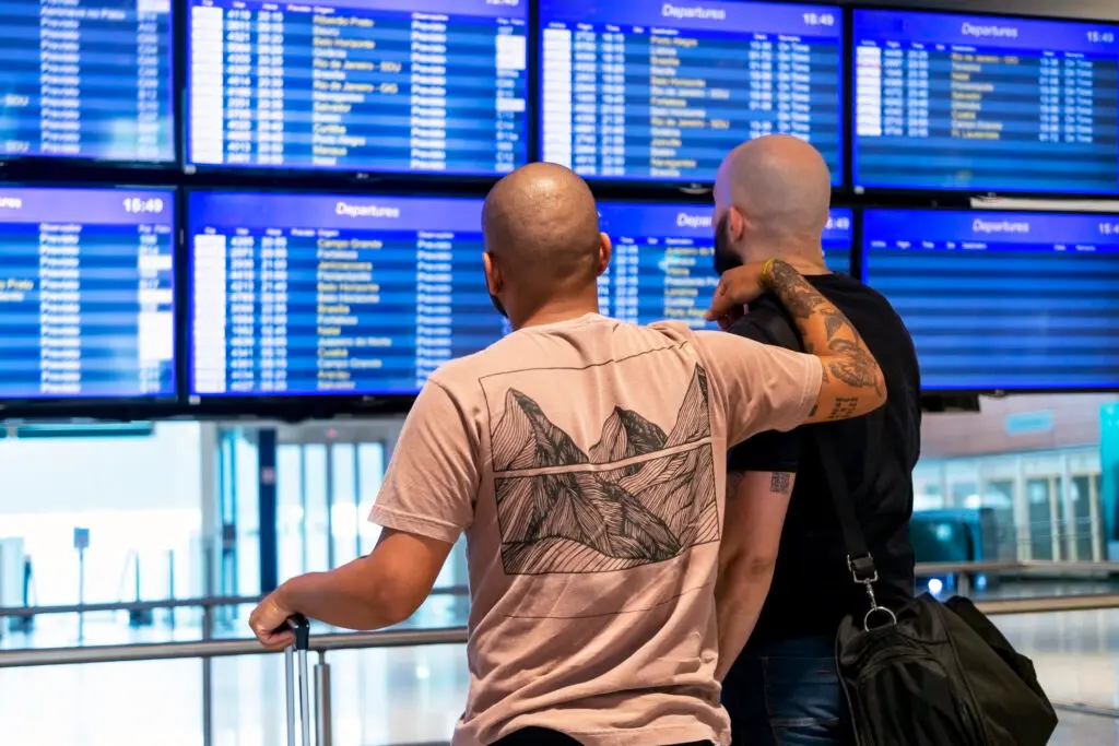 Two men view flight ETAs at an airport; LuxCitizenship's LGBTQIA+ clientele is showing a rising interest in EU citizenship by ancestry. 